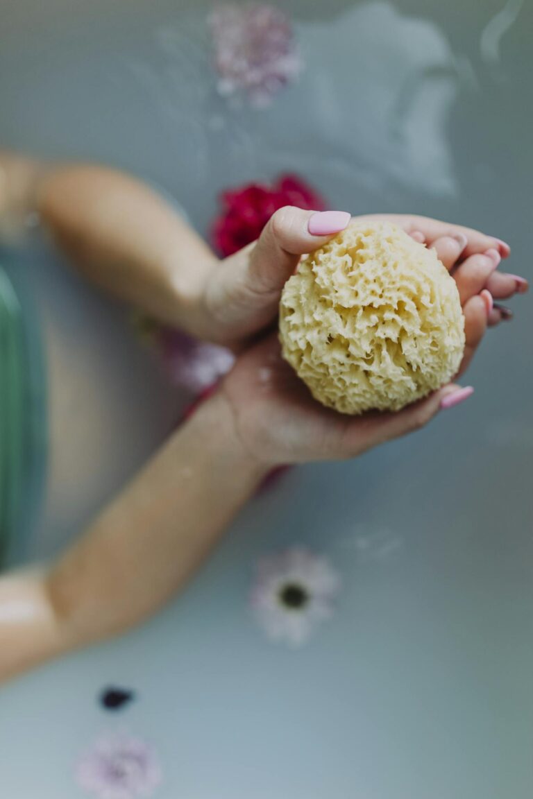 Hands holding a natural sea sponge over water surrounded by flowers for a relaxing spa experience.